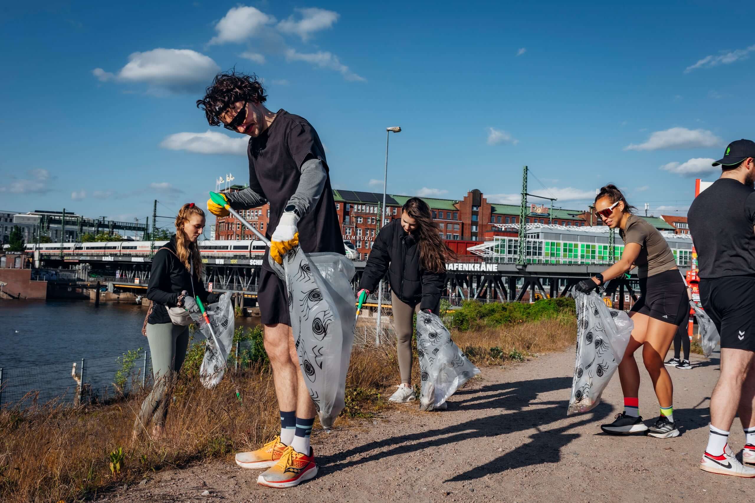 People picking up litter near a waterfront with buildings and a bridge in the background on a clear day.