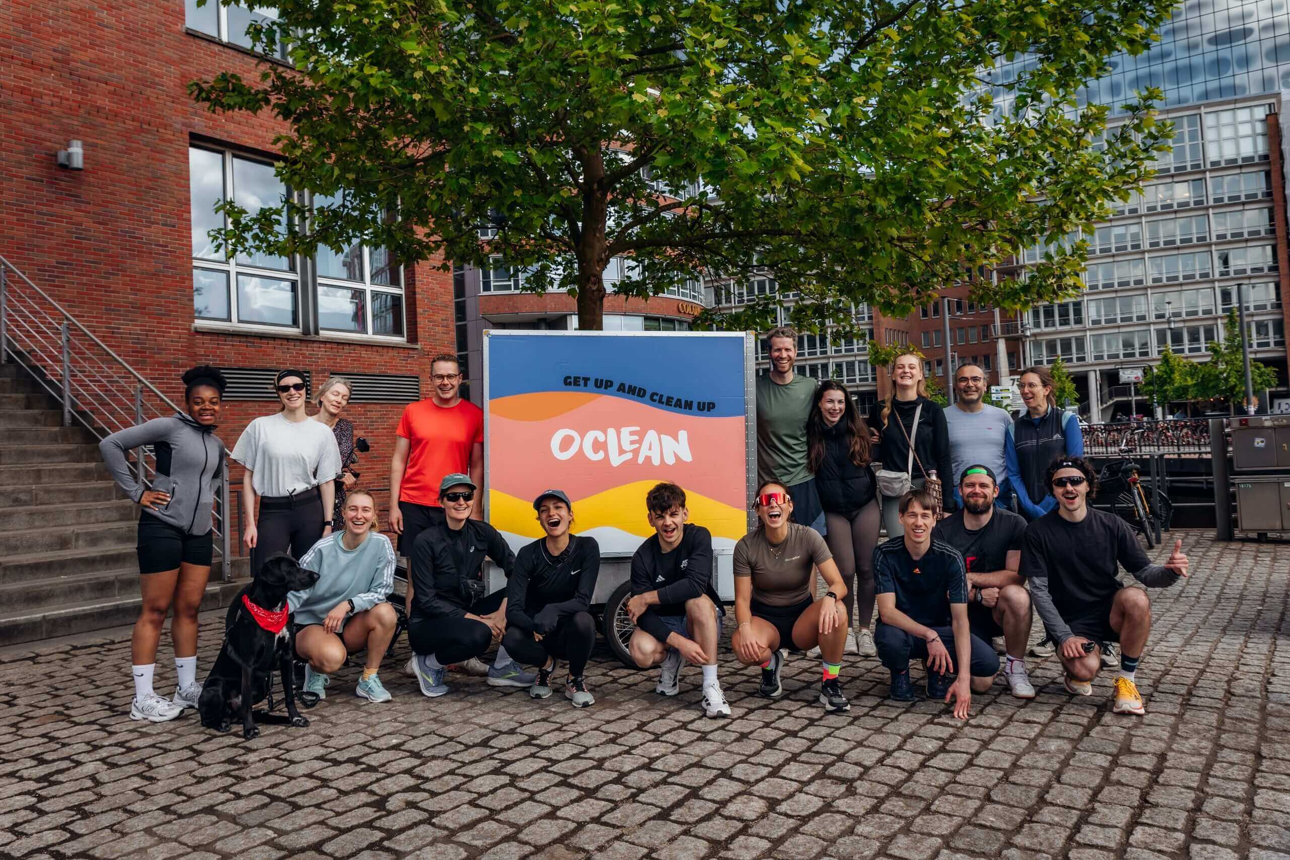 Große Gruppe lachender Teilnehmerinnen und Teilnehmer posiert gemeinsam mit einem Hund vor dem Oclean „Get Up and Clean Up"-Anhänger auf einem Kopfsteinpflasterplatz in Hamburg