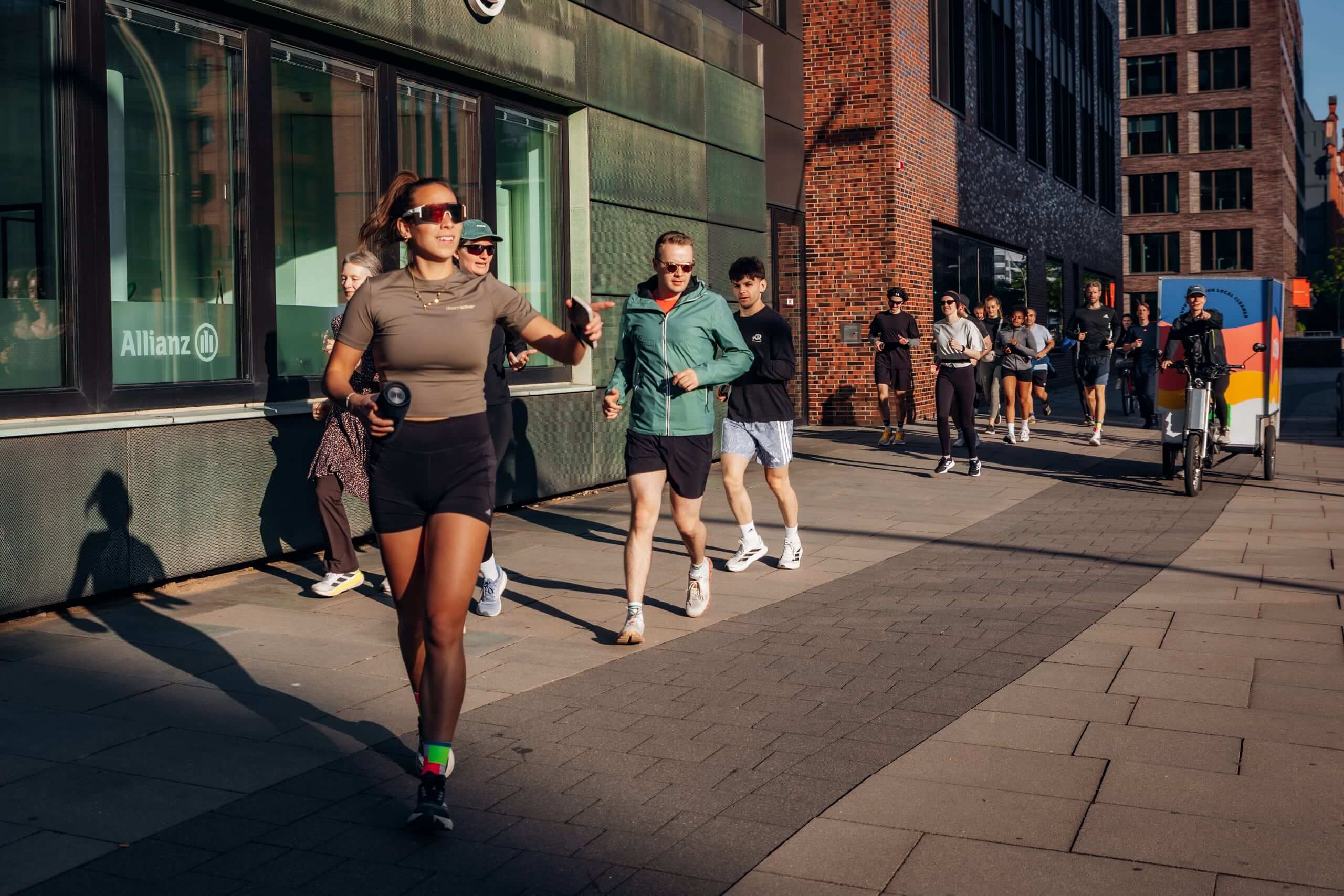 Gruppe von Läuferinnen und Läufern joggt bei Sonnenschein durch eine Hamburger Innenstadt, angeführt von einer Frau mit Sonnenbrille