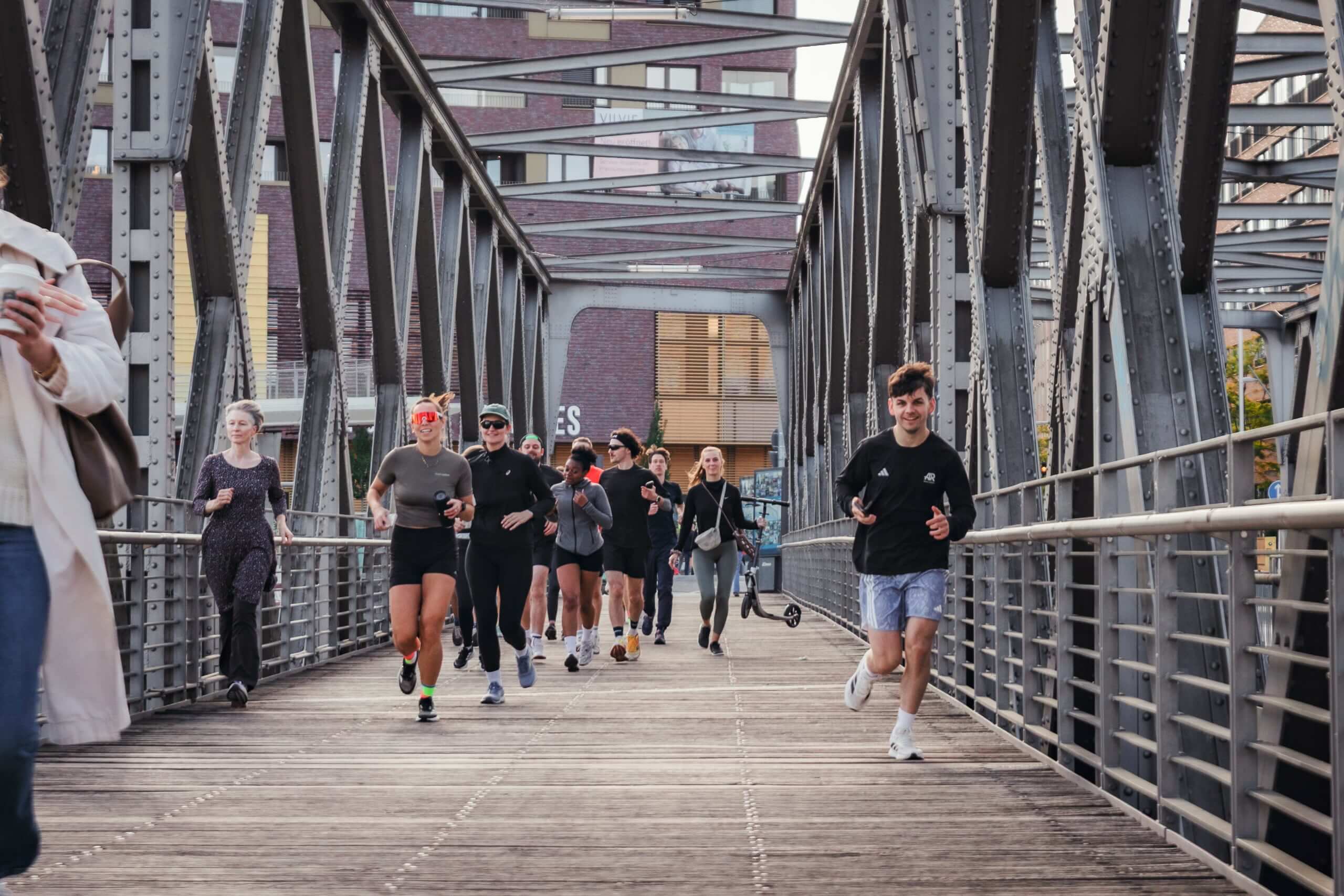 A group of people is jogging and walking on a metal bridge. They are dressed in athletic wear, and the cityscape is visible in the background.