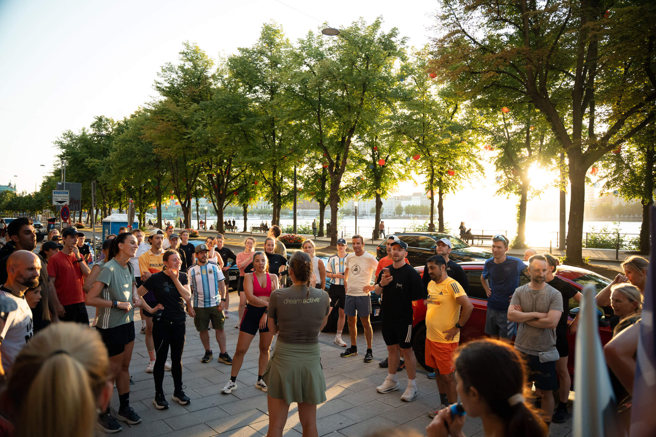 A group of people gathered outdoors under trees, listening to a speaker in sportswear, with sunlight filtering through the leaves. 1Komma5Grad