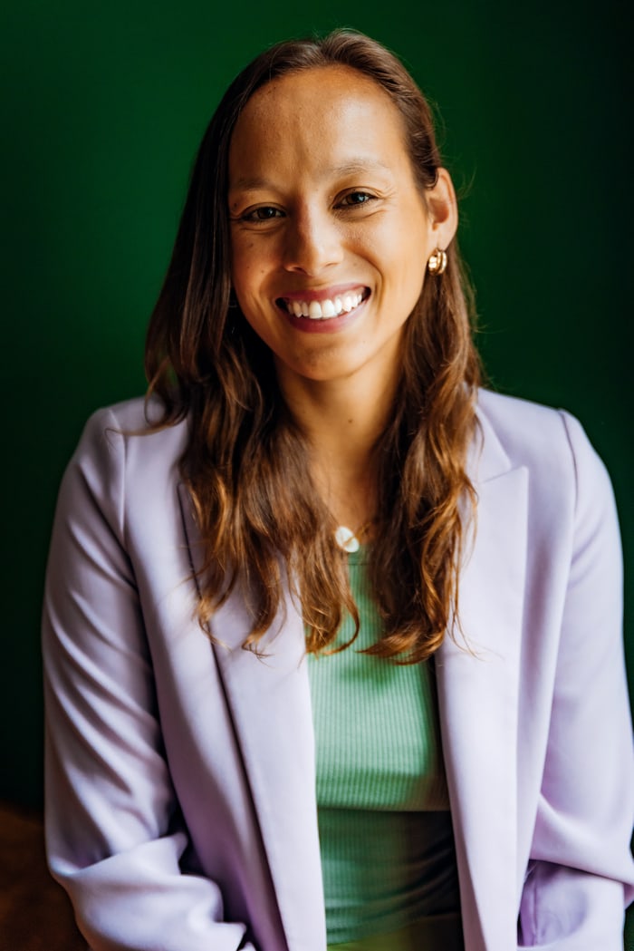 Porträt einer lächelnden Frau im lilafarbenen Blazer vor dunkelgrünem Hintergrund