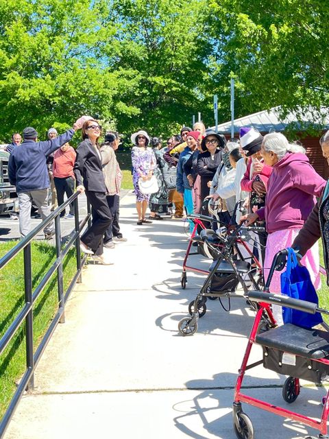 A group of seniors, some with walkers, stand and sit outdoors on a sunny day, surrounded by green trees and a clear sky.