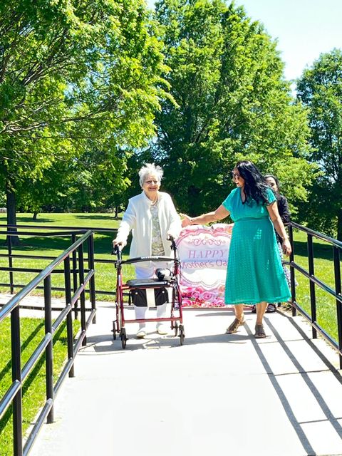 An elderly woman using a walker is assisted by a younger woman on a sunny park path, holding a "Happy Birthday" banner.