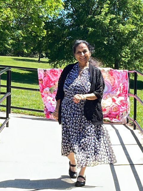 Woman in floral dress and black shawl smiles, standing on a sunny path with a vibrant floral backdrop and trees in the background.