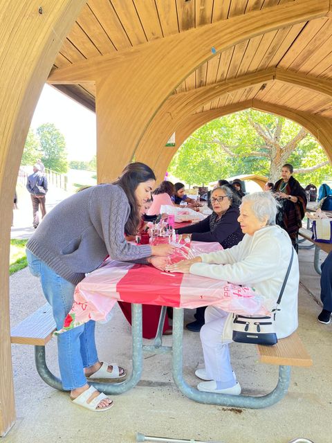 People gathered at a picnic table under a wooden pavilion, engaged in conversation, with trees and sunlight in the background.