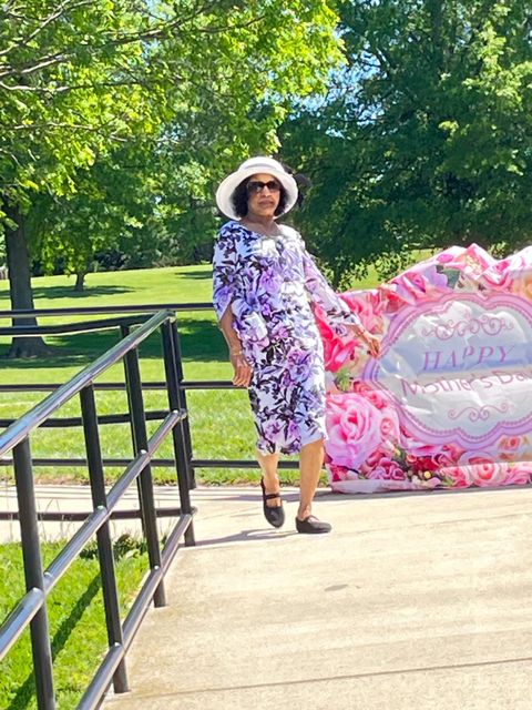 Woman in a floral dress and sun hat walks on a path with a "Happy Mother's Day" sign and greenery in the background.