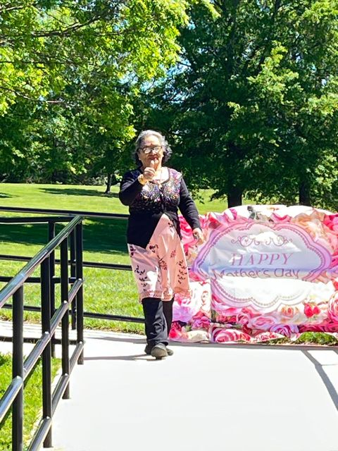 Elderly woman walking on a path, holding a drink, next to a "Happy Mother's Day" sign with floral decorations. Sunny day with green trees.