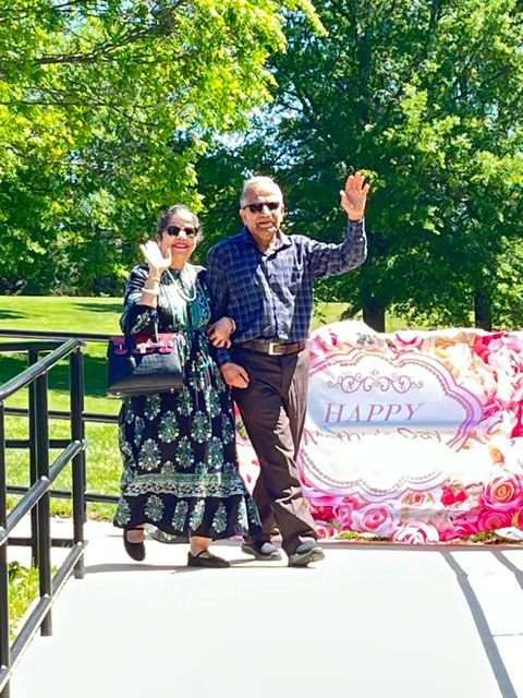 A smiling couple waves while walking on a sunny path, with a floral banner displaying "Happy" in the background.