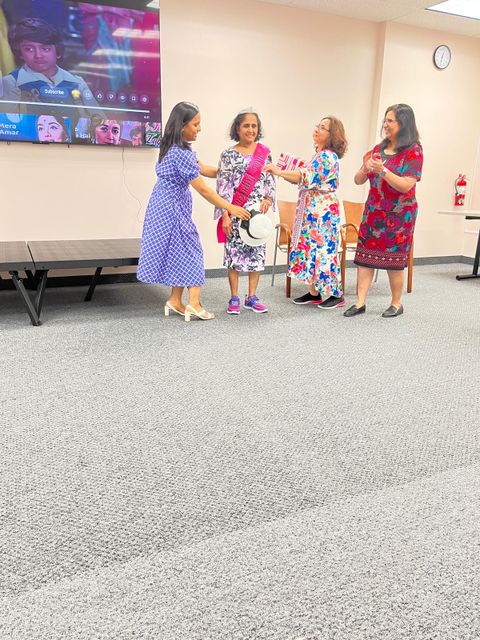 Four women in colorful dresses stand together in a room. One holds a sash and gift, while others smile. A screen displays a video call behind them.