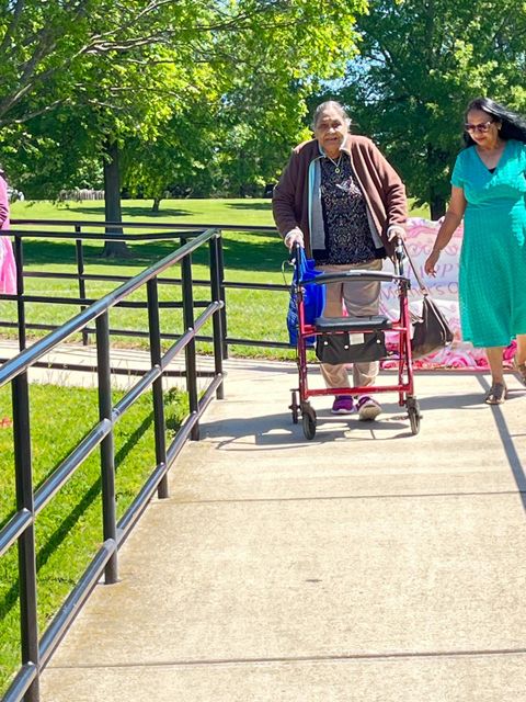 Elderly woman using a walker on a sunny path, accompanied by a woman in a green dress, with trees and grass in the background.