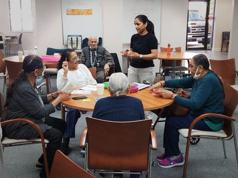 Shivangi Patel stands speaking to a group of participants seated around a table, engaged in conversation, in a community room setting.