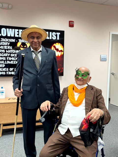 Two elderly men in suits, one with a cane and straw hat, the other in a mask and scarf, pose in a room with Halloween decorations.