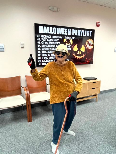 Person dressed in a Halloween costume with a cane, wearing sunglasses and a hat, in front of a "Halloween Playlist" screen.
