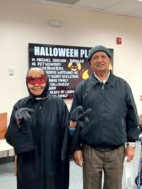 Two people in Halloween costumes, one wearing a red mask and cape, the other in a beanie and jacket, stand in front of a Halloween playlist sign.