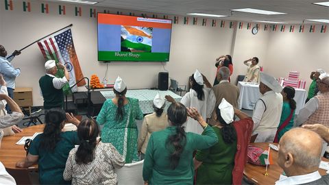 People in traditional attire saluting the Indian flag on a screen in a decorated room with Indian and American flags.