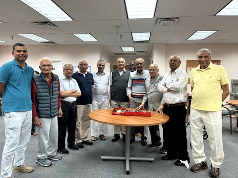 A group of elderly men stand around a table with a red cloth in a brightly lit room, smiling at the camera.