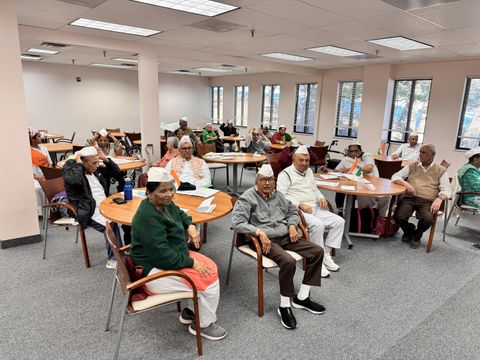 A group of elderly people wearing traditional caps sit at tables in a well-lit room, attentively facing the front.