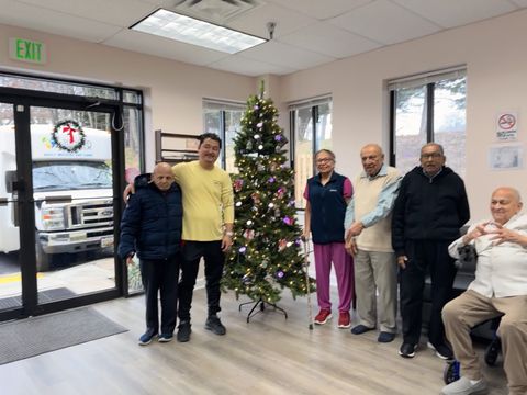 A group of six people stands inside a room decorated with a Christmas tree. A bus is visible outside the window.