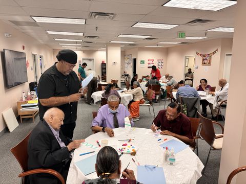 A group of elderly individuals participating in an art class, painting and drawing at tables in a brightly lit community room.