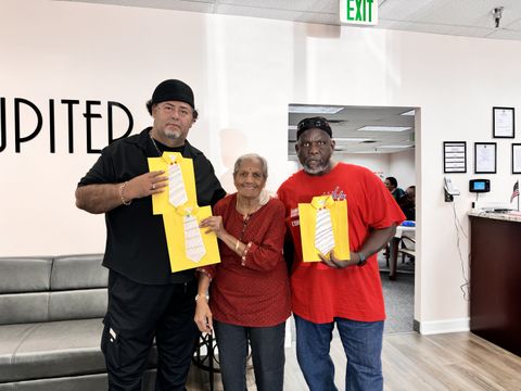 Three people standing together indoors, each holding a yellow paper with a drawing of a tie. They are smiling at the camera.