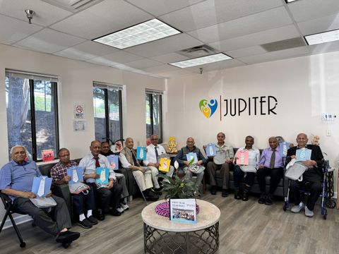 A group of elderly men sitting in a room, each holding a gift bag, with "Jupiter" logo on the wall behind them.