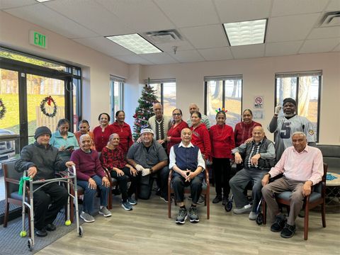 A group of elderly people and caregivers posing in a room with a Christmas tree, wearing festive clothing and smiling.