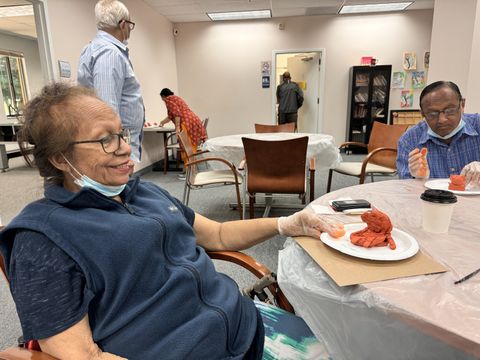 Elderly individuals engaged in a crafts activity, sculpting with clay at a community center, wearing gloves and masks, seated around tables.