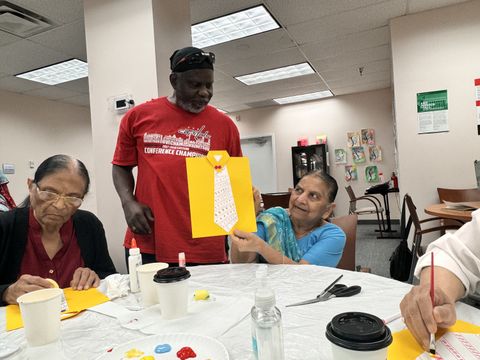 A woman shows a paper craft shirt to a man in a red shirt, while another woman works on crafts at a table with art supplies.