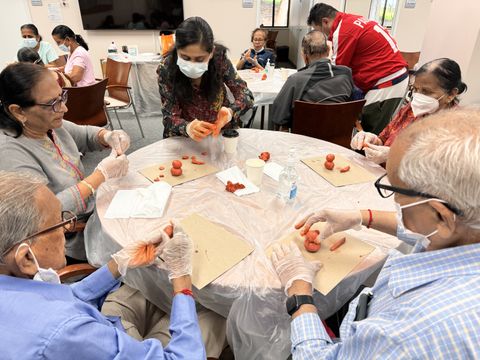 A group of elderly people and a caregiver, all wearing masks and gloves, sit around a table making crafts with red clay.