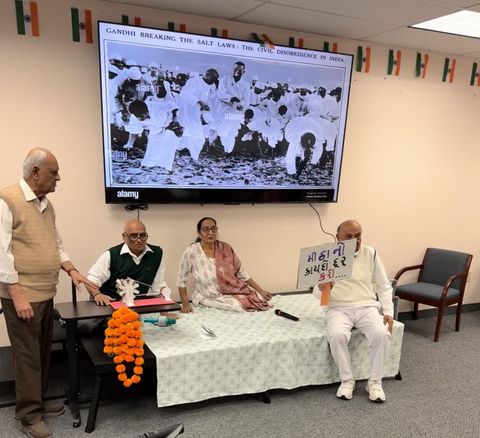 Four elderly individuals in a room, one holding a sign. Behind them is a large photo of Gandhi leading a protest. Indian flags decorate the walls.