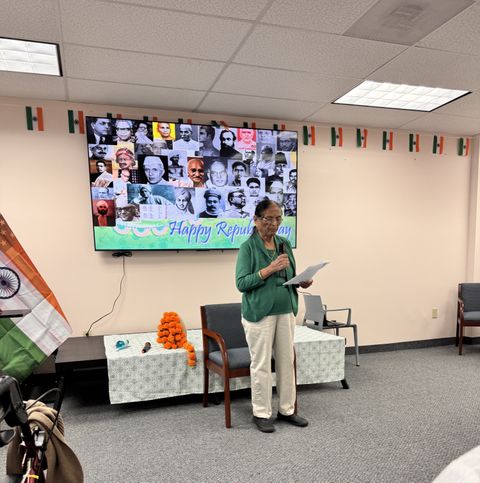 An elderly person speaks at a Republic Day event, with a collage of historical figures on a screen and Indian flags decorating the room.