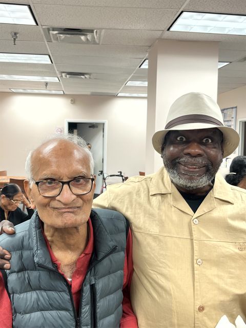 Two elderly men smiling, one wearing glasses and a puffer vest, the other in a beige shirt and hat, standing indoors with others in the background.