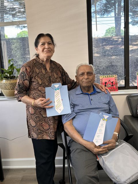Elderly couple smiling, holding handmade cards. The man is seated, the woman stands beside him. Indoor setting with a window and plants.