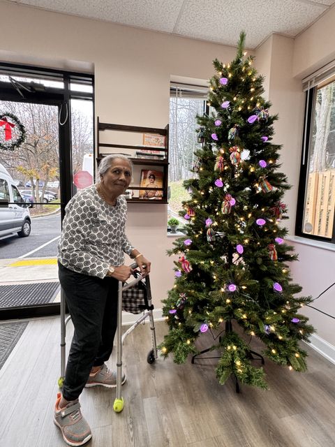 Elderly woman with a walker stands beside a decorated Christmas tree in a room with large windows and a bookshelf.