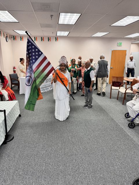 A woman in traditional attire holds the American and Indian flags in a room decorated with flags and people in the background.