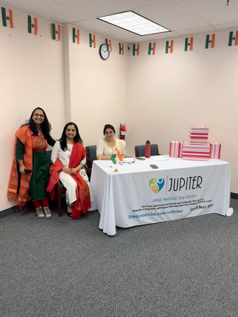 Three people smiling in a decorated room with a table covered by a "Jupiter" cloth, stacked gift boxes, and Indian flags on the walls.