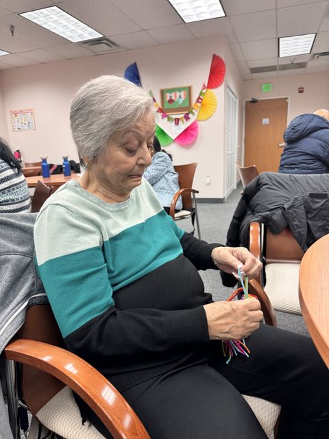 Elderly woman in a teal and black sweater sitting in a chair, sorting colorful pipe cleaners in a decorated room.