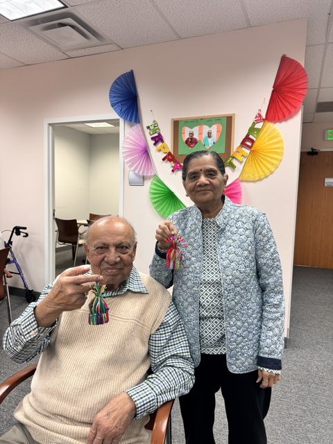 Two elderly people smiling, holding colorful tassels. Behind them, a "Happy Birthday" banner and colorful decorations adorn the wall.