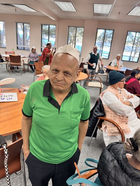Elderly man in a green shirt and paper hat stands in a room with others seated around tables, engaging in activities.