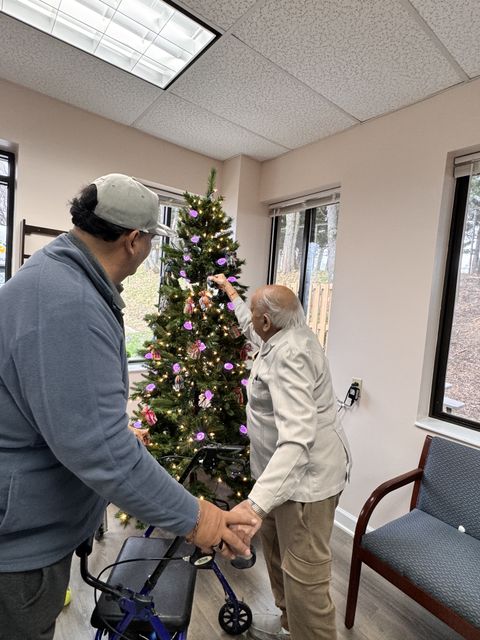 Two people decorating a Christmas tree in a room, one using a walker. The tree is adorned with lights and colorful ornaments.
