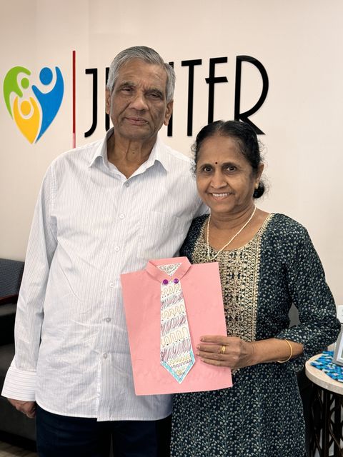 Elderly couple smiling, woman holding a pink paper shirt with a colorful tie drawing, standing in an office with a logo on the wall.