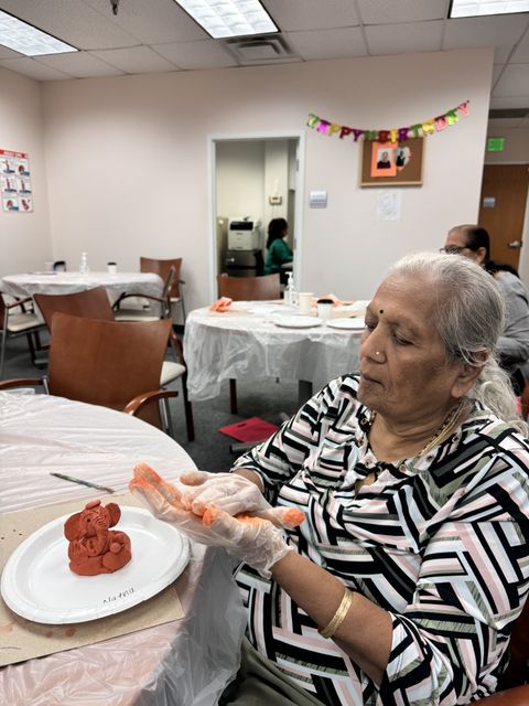 An elderly woman molds clay at a table in a decorated room, with a "Happy Birthday" banner in the background.