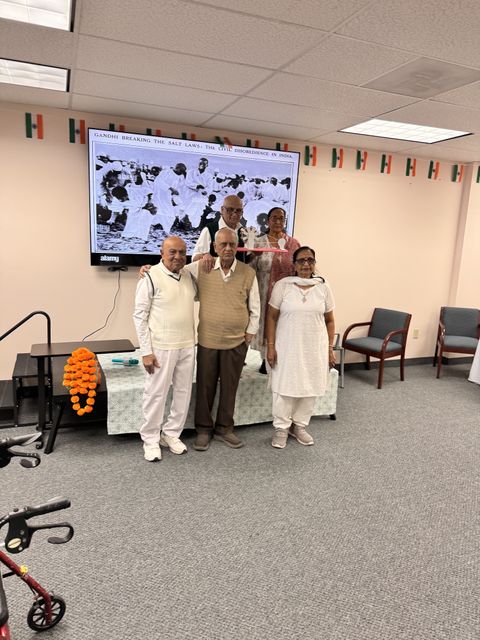 Five people stand together in a room decorated with Indian flags, in front of a screen displaying an image of Gandhi.