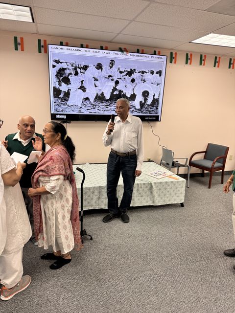 A man speaks into a microphone in a room with people gathered, a historic photo of Gandhi's Salt March displayed on a screen behind him.