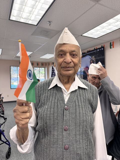 Elderly man in a white cap holds an Indian flag indoors, with another person in the background adjusting a similar cap.
