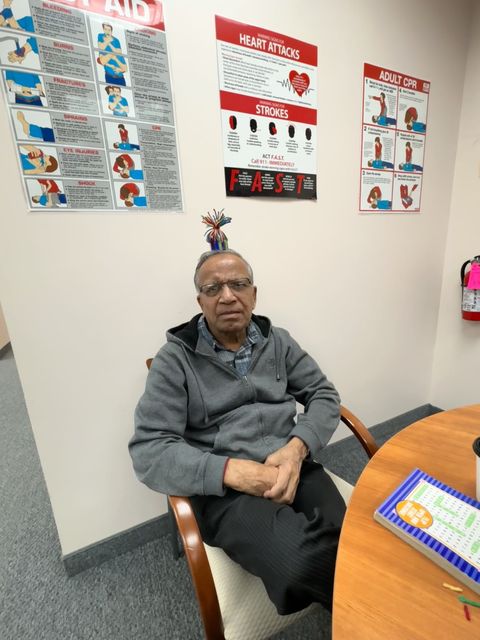 Man sitting in a chair, wearing glasses, a hoodie, and a funny hat, in a room with medical posters on the wall.