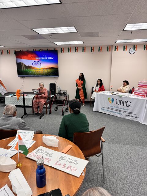 People in a room decorated with Indian flags, celebrating Republic Day. A woman speaks at a podium, others are seated at tables.