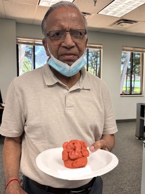 Elderly person holding a paper plate with a small clay sculpture resembling an elephant, wearing a mask under his chin, standing indoors.
