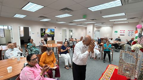 A group of elderly people in a community room participate in a prayer session, with one man standing and others seated, hands folded in reverence.